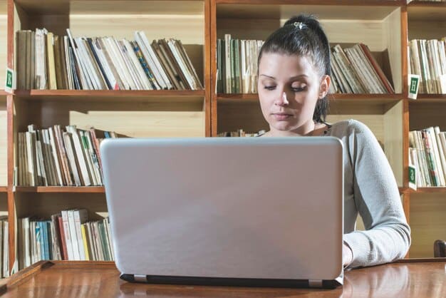 A student using a laptop in a library, surrounded by books and research materials. The focus is on the laptop screen, which displays the FAFSA application form.