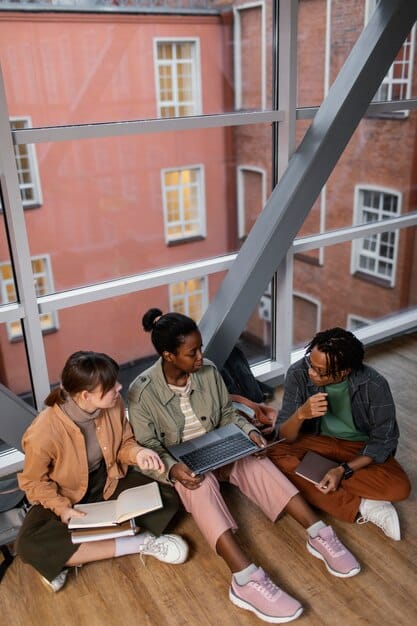A diverse group of college students engaged in studying, some using laptops, others reading textbooks, in a modern university library setting, illustrating accessibility through grants.
