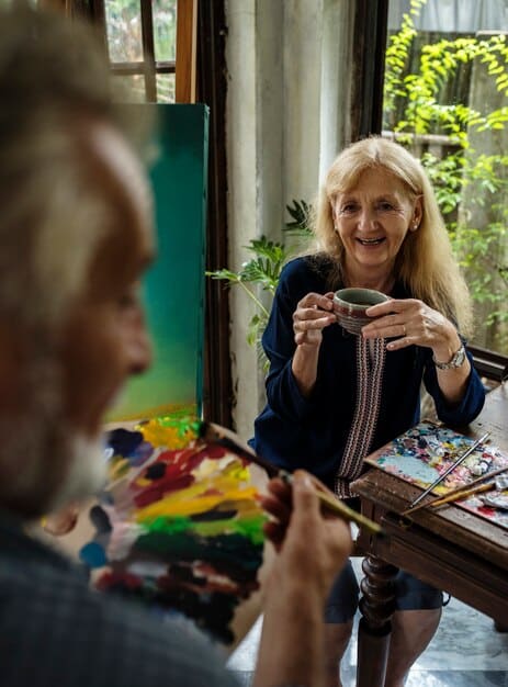 An elderly woman smiling while engaged in a creative art activity with a caregiver in a bright common area, showcasing the importance of personalized activities and respectful interaction.