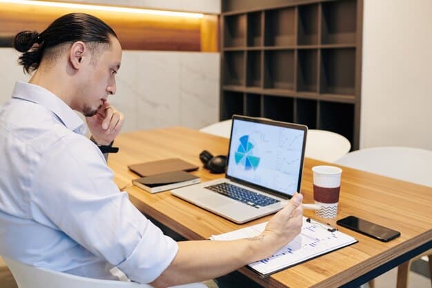 A person sitting at a desk, looking at a laptop screen filled with charts and data related to healthcare costs and plans, with a thoughtful expression. The background is a modern home office.